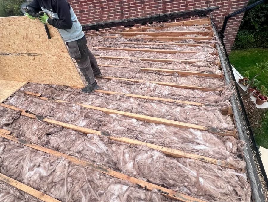Flat roof stripped back to the joists with new insulation between them, a roofer bringing up the first deck boards